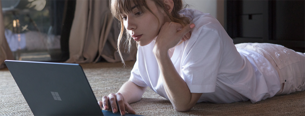 A woman at home lying down on her carpet using her open Microsoft Surface Laptop